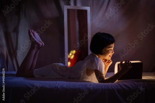 Bella niña asiática leyendo libro luminoso en la cama