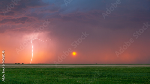 Lightning bolt strikes at sunset with the sun seen heavily filtered by the rain falling in the distance. Dark storm clouds can be seen overhead.