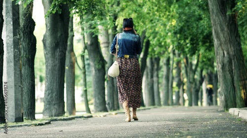 Woman walks along sidewalk on warm summer day. Courier cyclist with yellow backpack   seen in distance. Trees with fresh green foliage grow around. City street during day. Beginning of June.