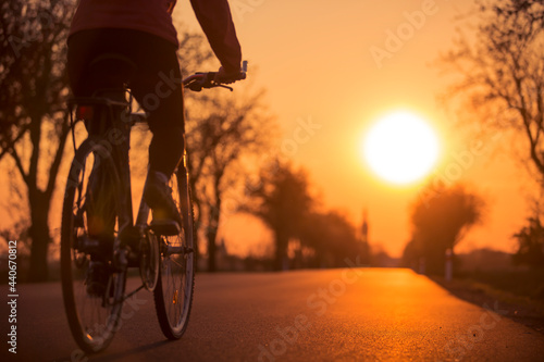 Blurred silhouette of young woman cycling on road to village during sunset. Tree alley on edges of asphalt road.