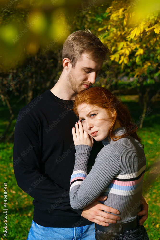 a young couple of Caucasian ethnicity, a man and a woman with red hair are standing in an embrace in a park on a sunny day against the background of trees a young couple of Caucasian ethnicity, a man and a woman with red hair are standing in an embrace in a park on a sunny day against the background of trees