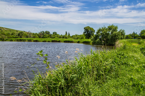 The shore of the lake is overgrown with grass and shrubs. Windy weather causes ripples on the water. The blue sky is covered with light clouds.