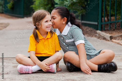 Two school girls sitting outside one child whispering secrets to other kid photo