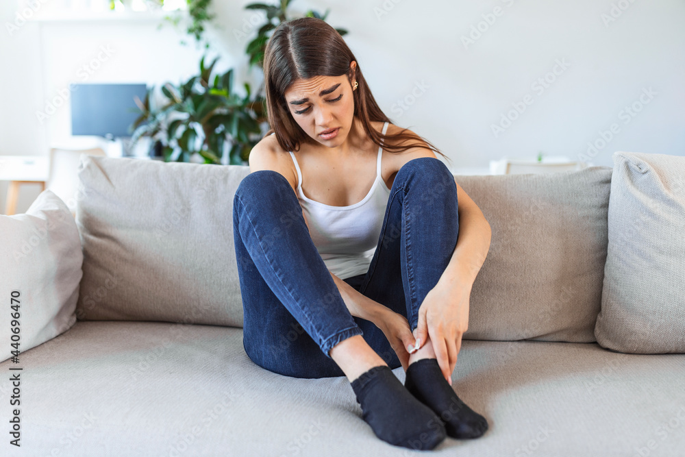 Hands massaging swollen foot while sitting on sofa during the day at home. Photo of Young Caucasian woman suffering from pain in leg. Woman massaging her legs after all day at work in office Hands massaging swollen foot while sitting on sofa during the day at home. Photo of Young Caucasian woman suffering from pain in leg. Woman massaging her legs after all day at work in office