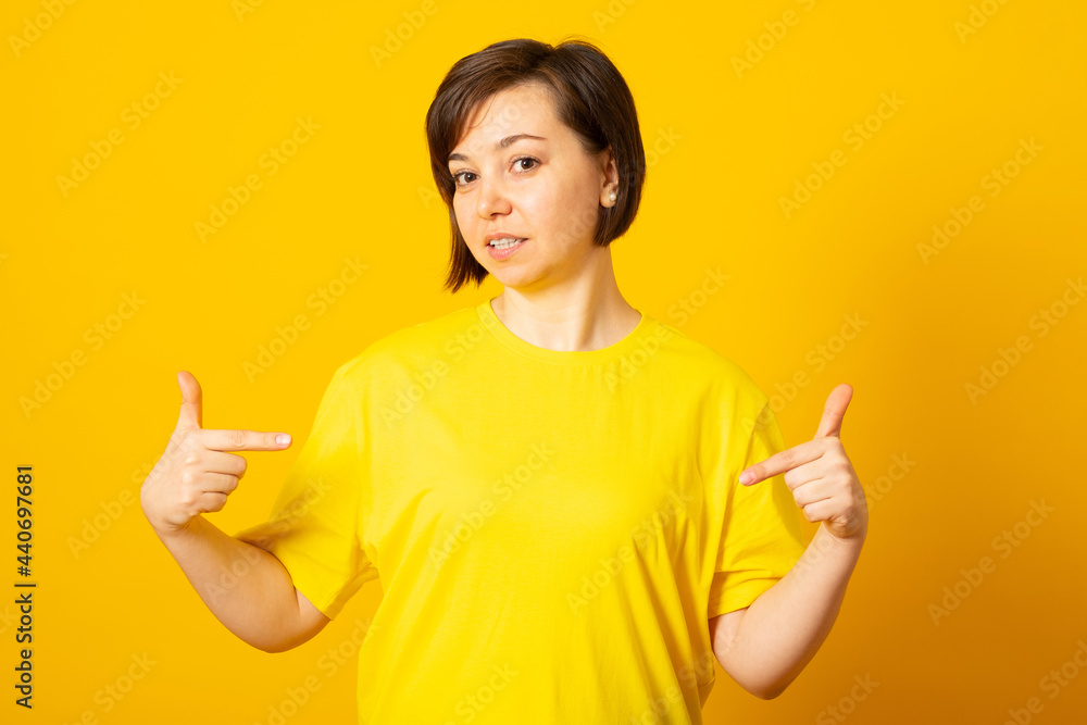 T-shirt design and advertising concept. Style and fashion. Indoor shot of cheerful smiling young woman pointing index finger at copy space on her blank white top. T-shirt design and advertising concept. Style and fashion. Indoor shot of cheerful smiling young woman pointing index finger at copy space on her blank white top.