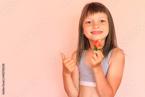 The concept of the benefits of early vitamin berries. Smiling eight-year-old girl holds a large strawberry, on a light background,in the other hand, the finger is raised up, close-up