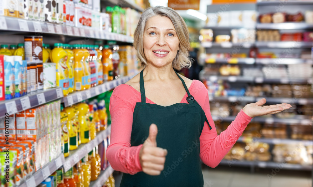Smiling confident mature saleswoman giving thumbs up, recommending quality food products in supermarket