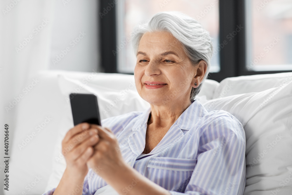 old age, technology and people concept - happy smiling senior woman in pajamas using smartphone sitting in bed at home bedroom old age, technology and people concept - happy smiling senior woman in pajamas using smartphone sitting in bed at home bedroom
