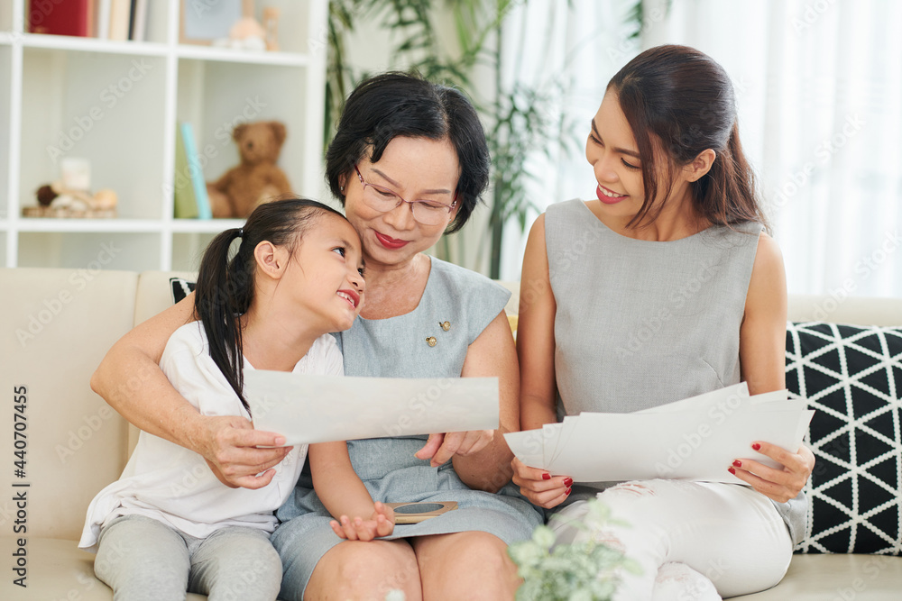 Adorable creative little girl showing her drawings to mother and grandmother when they are sitting on sofa at home Adorable creative little girl showing her drawings to mother and grandmother when they are sitting on sofa at home