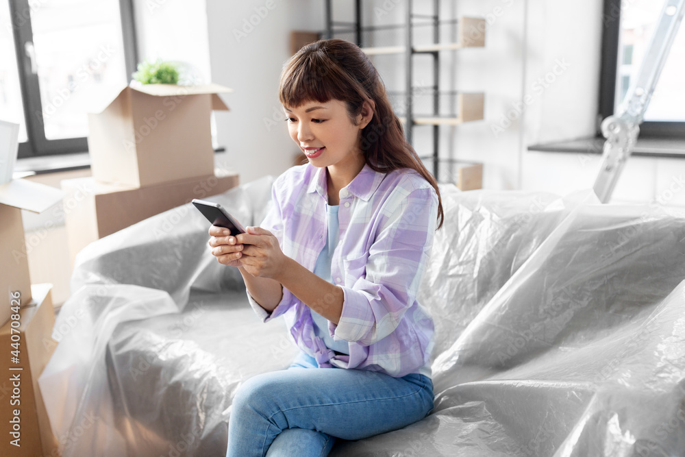 moving, people and real estate concept - happy smiling asian woman with smartphone and boxes at new home moving, people and real estate concept - happy smiling asian woman with smartphone and boxes at new home