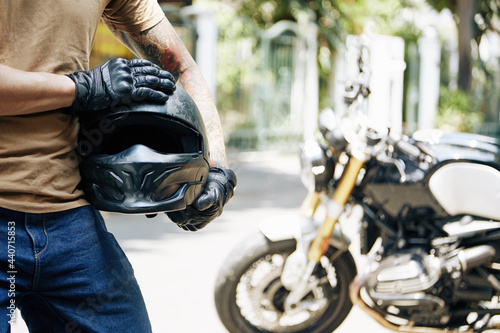 Cropped image of motorcyclist in leather gloves holding his helmet, his bike is in background