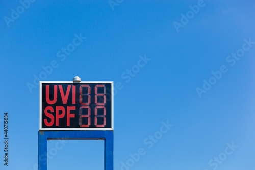 Electronic display with UV radiation warning and info about recommended SPF with red letters, bright blue summer sky in background