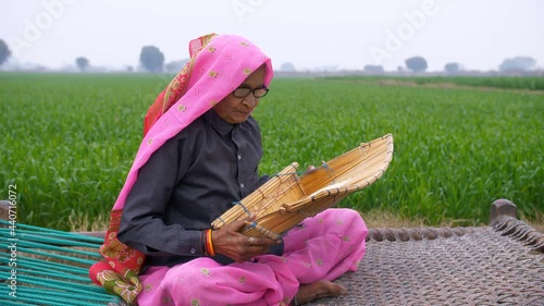 An old Indian woman from a village cleaning rice using a bamboo rice-cleaner. An elderly lady of an Indian village cleaning rice using traditional rice cleaner while sitting near a field on a chaarpai photo