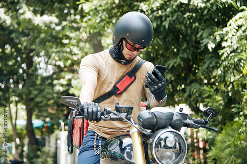Deliveryman in helmet sitting on motorcycle and checking address of customer via application on smartphone