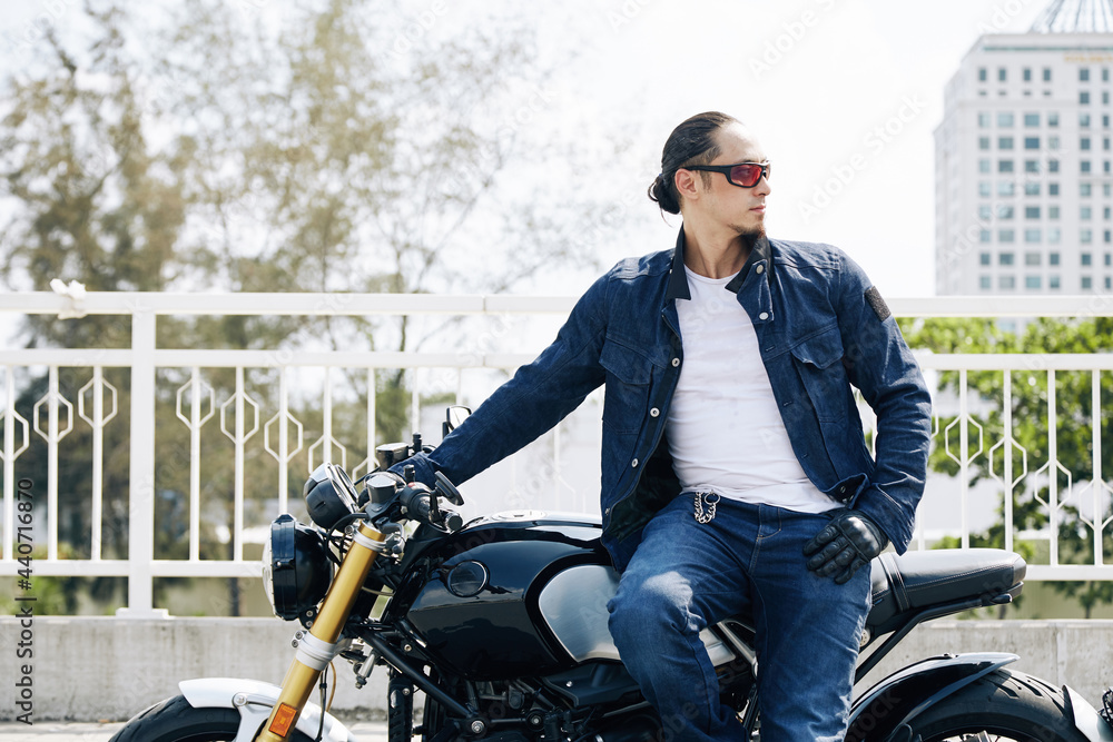 Portrait of cool stylish man in sunglasses sitting on motorcycle and looking away