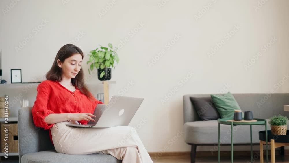 A beautiful young businesswoman in a bright red shirt is typing on a laptop while sitting in a comfortable armchair at the office A beautiful young businesswoman in a bright red shirt is typing on a laptop while sitting in a comfortable armchair at the office