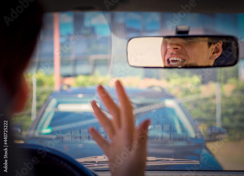 Driver sitting in his car behind the wheel shaking his hand threatens another car ahead.Negative emotion reflected in the mirror.