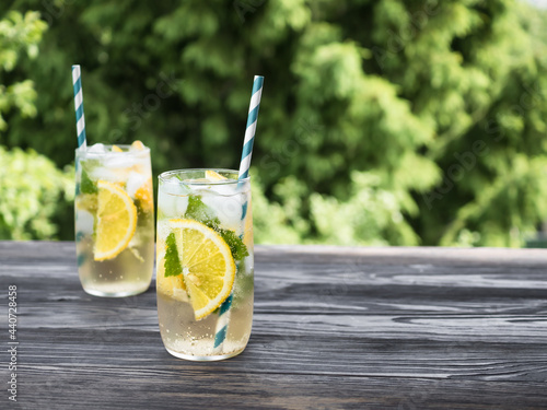 Cold carbonated lemonade with ice, lemon and currant leaves in a transparent glass. Summer drink stands on a wooden table in the garden. Background - fir branches