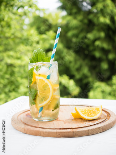 Glasses of cold lemonade with ice and currant leaves on a wooden tray. Summer drink in the garden. Green branches in the background.