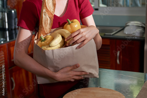 Close-up of unrecognizable woman standing at counter and embracing paper bag with fresh products such as bell pepper, bananas, baguette