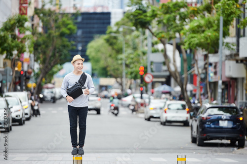 Positive young Asian guy in sun hat standing on small street pole against high traffic and drinking coffee