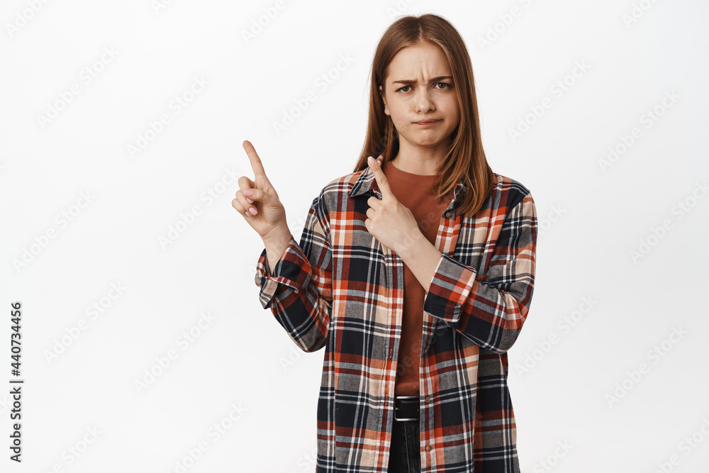 Skeptical girl furrow eyebrows, smirk and squint upset, pointing at upper left corner upset, dislike smth bad, being unsure, express her disbelief, white background Skeptical girl furrow eyebrows, smirk and squint upset, pointing at upper left corner upset, dislike smth bad, being unsure, express her disbelief, white background