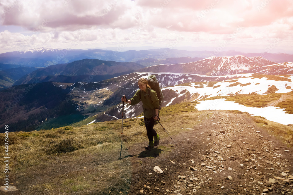 Young smiling girl climbs the mountains with trekking poles and a backpack in early spring, on the background there is a beautiful view with snowy mountains. Traveler goes hiking in the national park.