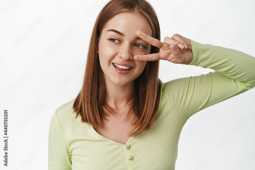 Close up young blond woman in green stylish bloud, show peace v-sign gesture near eye, smiling and looking left at promo beauty text, standing against white background Close up young blond woman in green stylish bloud, show peace v-sign gesture near eye, smiling and looking left at promo beauty text, standing against white background