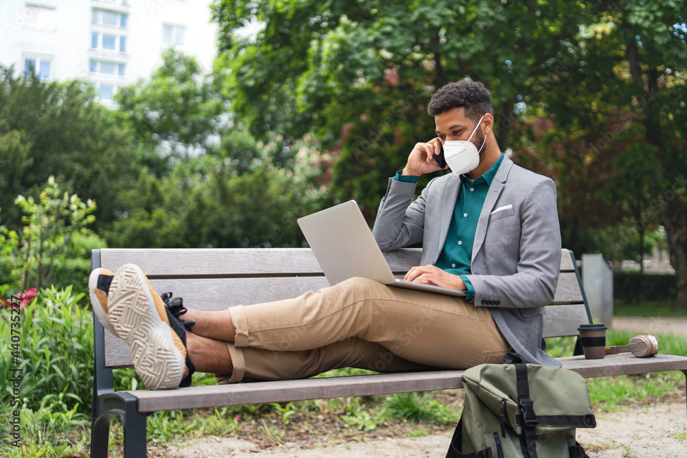Fototapeta premium Young man with respirator using laptop and smartphone outdoors in city, covid-19 concept.