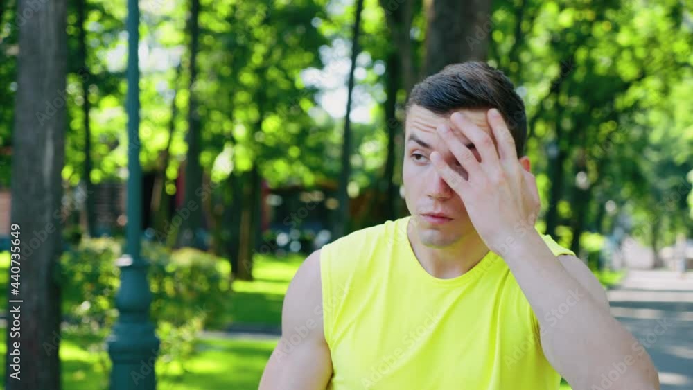 Young muscular man wearing yellow sports shirt walking in park and breathing hard, drinking water from bottle, sweat on his face. Tracking shot athlete after workout outside. Concept of fitness Young muscular man wearing yellow sports shirt walking in park and breathing hard, drinking water from bottle, sweat on his face. Tracking shot athlete after workout outside. Concept of fitness