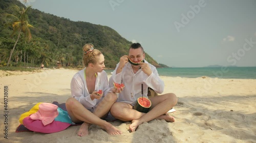 The cheerful love couple holding and eating slices of watermelon on tropical sand beach sea. Romantic lovers two people caucasian spend summer weekend in outdoor. Hat, backpack white shirt beachwear.