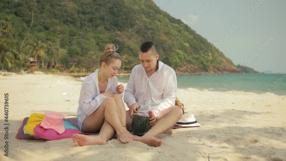 The cheerful love couple holding and eating slices of watermelon on tropical sand beach sea. Romantic lovers two people caucasian spend summer weekend in outdoor. Hat, backpack white shirt beachwear. The cheerful love couple holding and eating slices of watermelon on tropical sand beach sea. Romantic lovers two people caucasian spend summer weekend in outdoor. Hat, backpack white shirt beachwear.