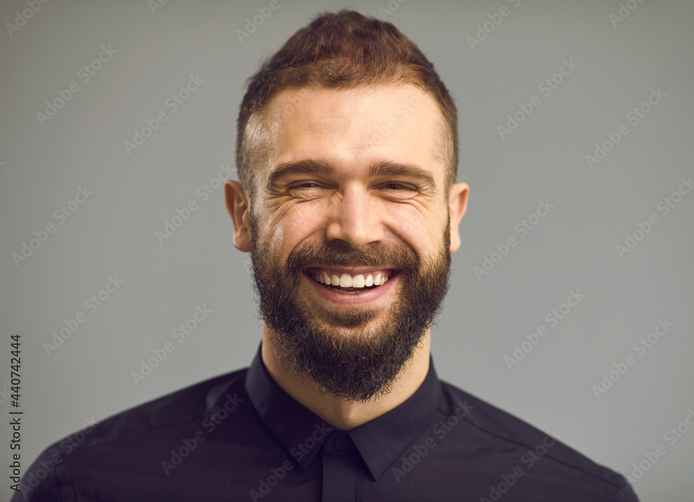 Obraz premium Close up portrait of a bearded man in a black shirt who is sincerely smiling and has wrinkles around his eyes. Concept of positive human emotions and people. Gray background. Banner. Place for text.