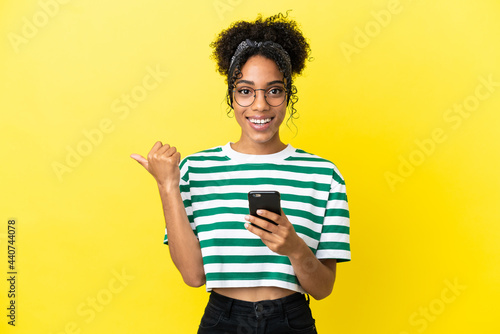 Young african american woman isolated on yellow background using mobile phone and pointing to the lateral
