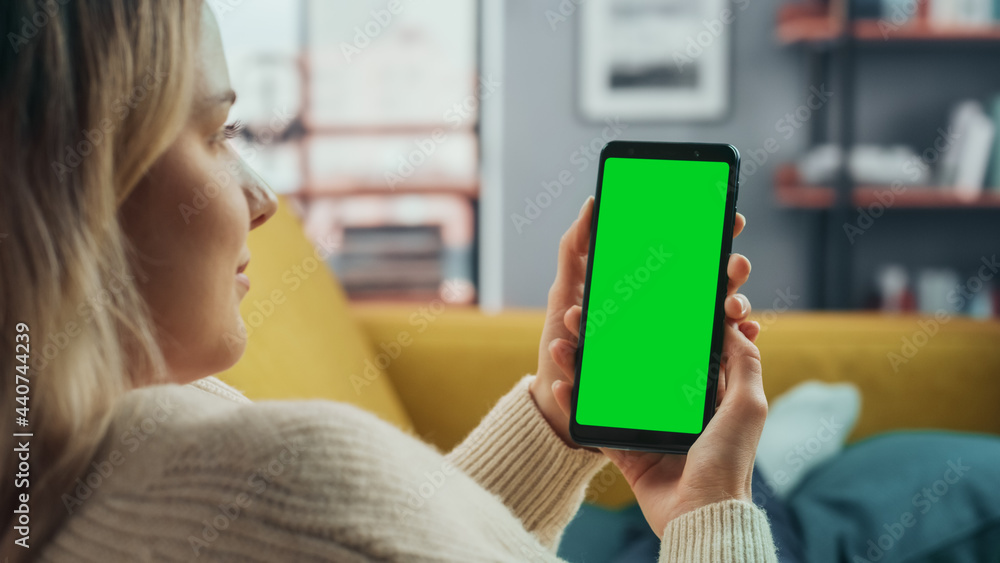Beautiful Authentic Female Using a Smartphone with Green Screen Mock Up Display at Home Living Room while Lying on a Couch Sofa. She's Browsing the Internet and Checking Videos on Social Networks.