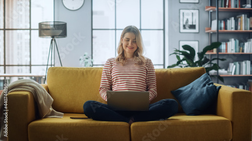Beautiful Caucasian Specialist Working on Laptop Computer at Stylish Home Living Room while Sitting on a Cozy Couch Sofa. Freelance Female Browsing Internet, Using Social Networks, Having Fun in Flat.