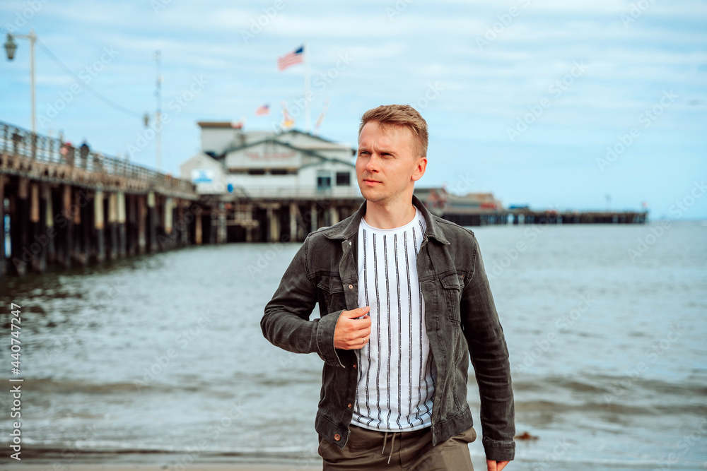A stylish young man in a denim jacket walks along the beach against the backdrop of the pier and the ocean in Santa Barbara, California, USA