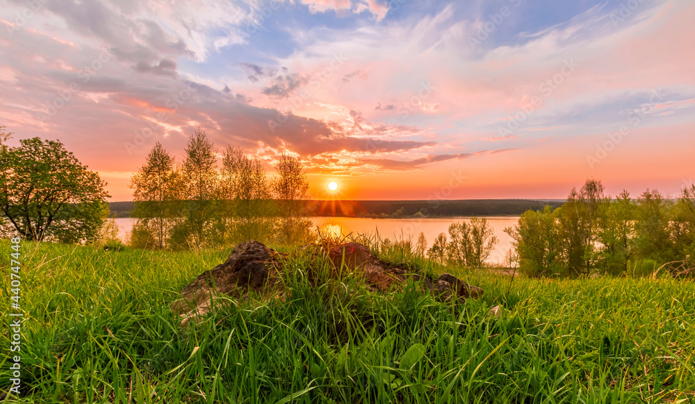 Scenic view at beautiful sunset on a shiny lake with old rough stone on the foreground, green grass, birch trees, golden sun rays, calm water ,nice cloudy sky on a background, spring landscape