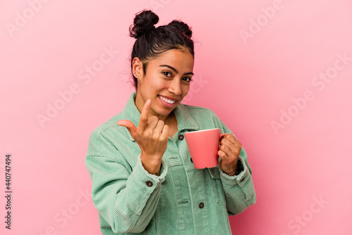 Tableau sur toile Young latin woman holding a mug isolated on pink background pointing with finger at you as if inviting come closer