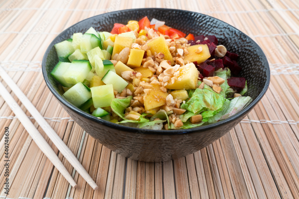 Vegetarian poke bowl in design black bowl with chopsticks below, on a reed pad background. Green cucumber, red pepper, peanuts, corn, beetroot on top of sushi rice. Healthy vegetarian food. Vegetarian poke bowl in design black bowl with chopsticks below, on a reed pad background. Green cucumber, red pepper, peanuts, corn, beetroot on top of sushi rice. Healthy vegetarian food.