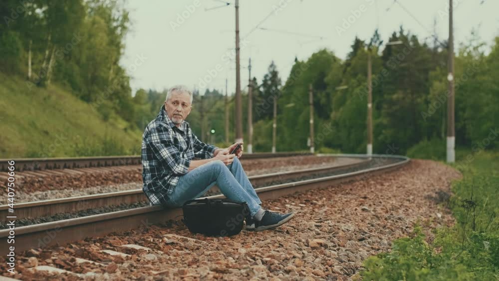 An elderly male tourist is sitting on the railway tracks, looking through information on the phone and waiting for the train. A middle-aged tourist waits for a train on the railway on a summer day. An elderly male tourist is sitting on the railway tracks, looking through information on the phone and waiting for the train. A middle-aged tourist waits for a train on the railway on a summer day.