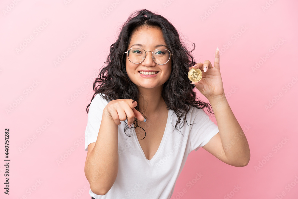 Young asian woman holding a Bitcoin isolated on pink background surprised and pointing front Young asian woman holding a Bitcoin isolated on pink background surprised and pointing front