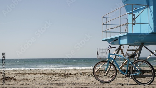Blue bicycle, cruiser bike by ocean beach, pacific coast, Oceanside California USA. Summertime vacations, sea shore. Vintage cycle on sand near lifeguard tower or watchtower hut. Sky and water waves.