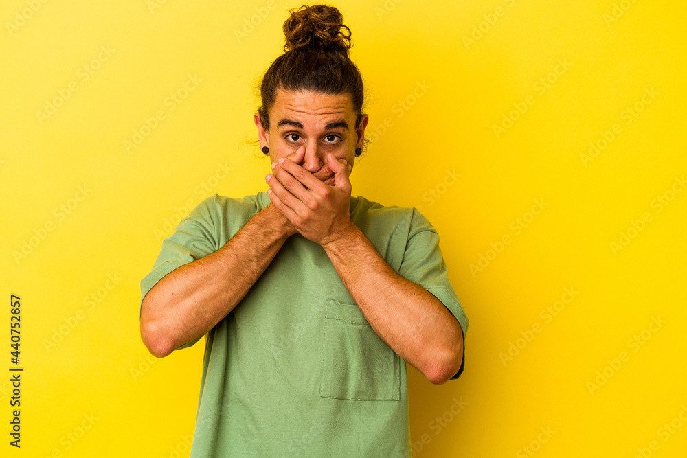 Young caucasian man with long hair isolated on yellow background covering mouth with hands looking worried. Young caucasian man with long hair isolated on yellow background covering mouth with hands looking worried.