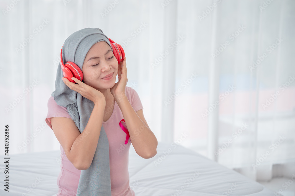 Asian woman wearing a hijab uses her smartphone and laptop in her bed. Attaching a pink ribbon represents recovery from a breast cancer patient. Breast cancer concept, cancer prevention concept. Asian woman wearing a hijab uses her smartphone and laptop in her bed. Attaching a pink ribbon represents recovery from a breast cancer patient. Breast cancer concept, cancer prevention concept.