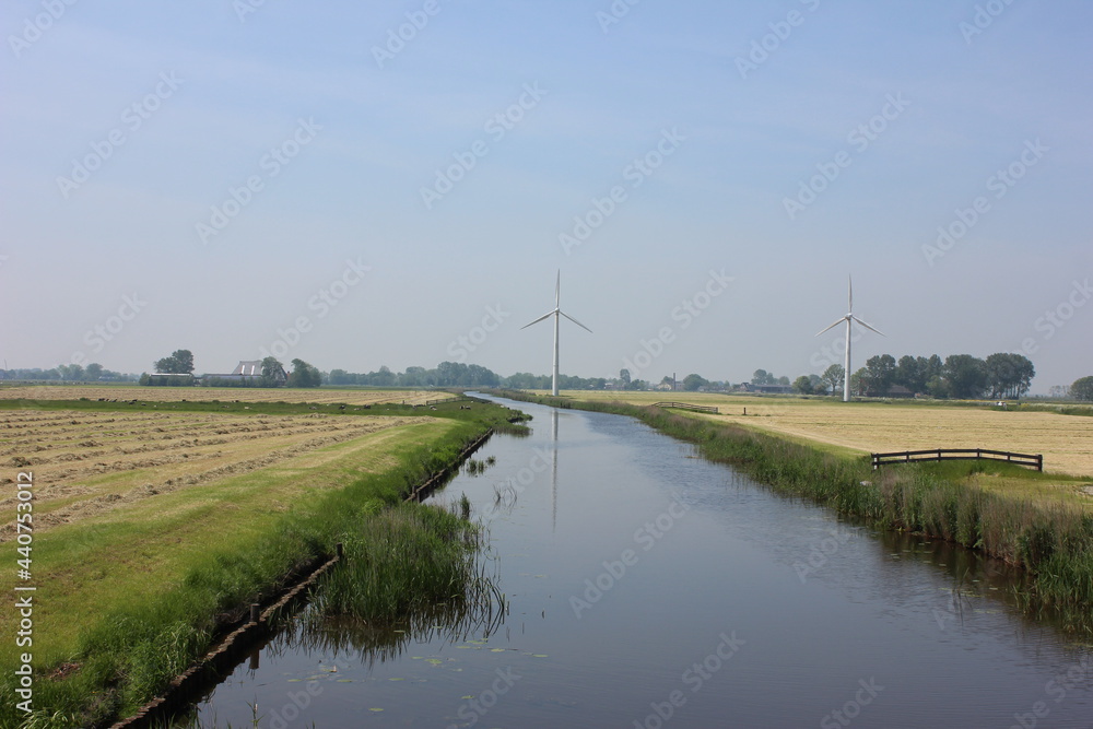 Wide view over a stream with meadows and wind turbines in the distance.