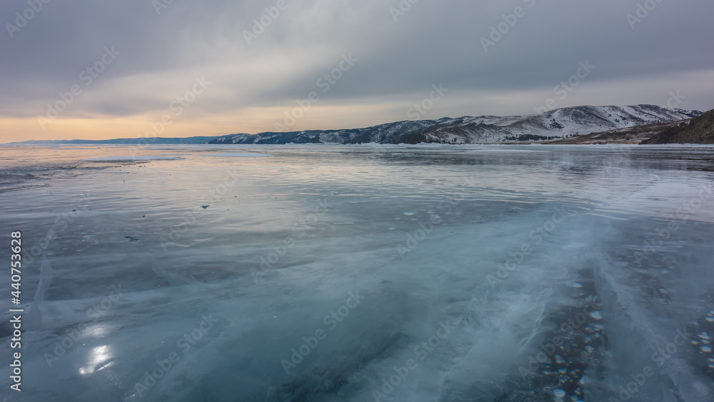 Sunset over a winter lake. The cloudy sky is highlighted in orange. Cracks on the smooth ice, bubbles of frozen methane gas. Reflections of the sun on the surface. Mountains on the horizon. Baikal