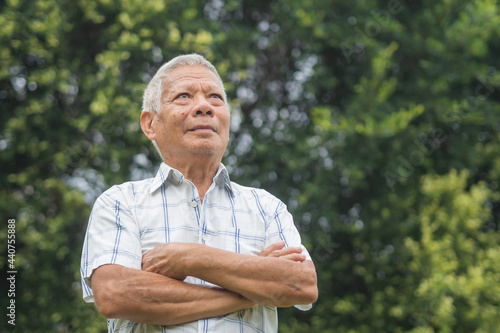Senior man arms crossed, smiling, and looking up while standing in a garden. Elderly Asian man happiness after retirement. Space for text. Aged people and relaxation concept
