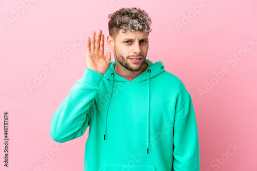 Young handsome caucasian man isolated on pink background listening to something by putting hand on the ear © luismolinero