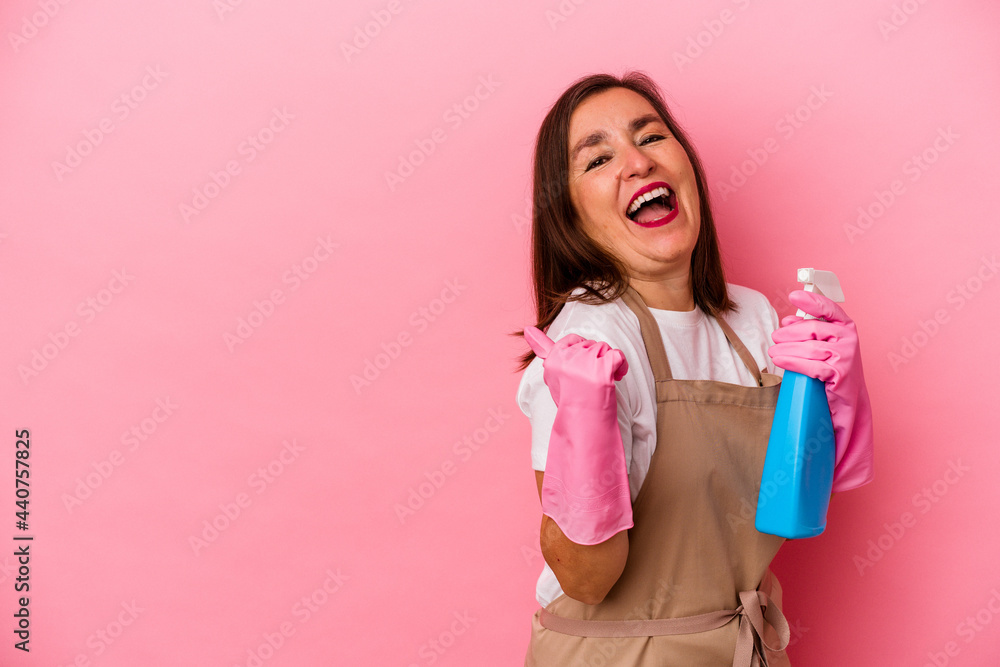 Middle age caucasian woman cleaning home isolated on pink background points with thumb finger away, laughing and carefree. Middle age caucasian woman cleaning home isolated on pink background points with thumb finger away, laughing and carefree.